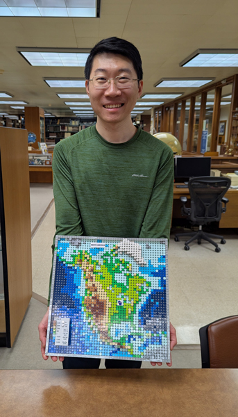 Photo of a young man in a library holding up a square map of North America constructed out of LEGO bricks.