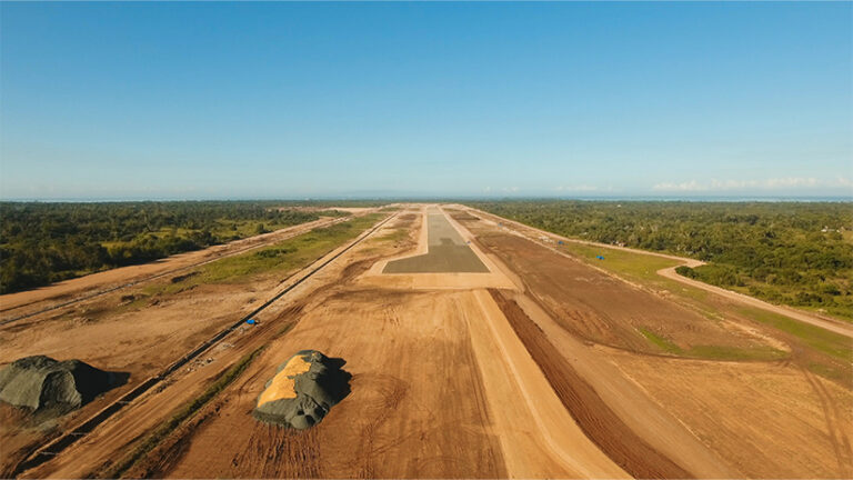 Landscape of a long dirt lot extending into the distance, with the beginnings of runway infrastructure and green vegetation on either side.
