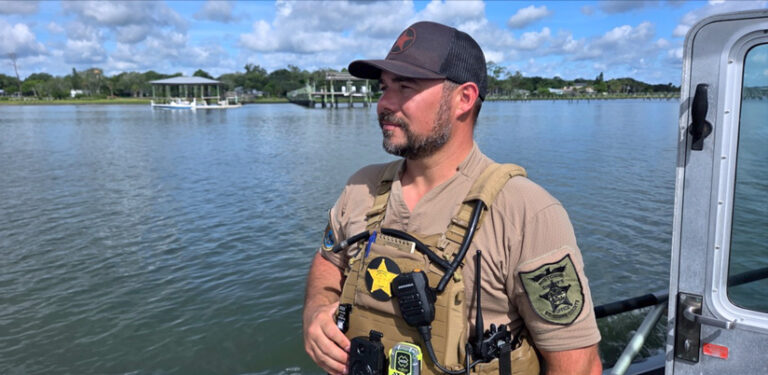 Photo of a man with a baseball cap and law enforcement uniform standing on the deck of a boat. A couple of structures can be seen on the coastline in the background.