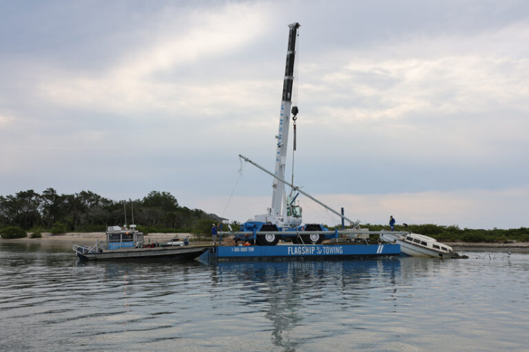 Photo of a boat being towed by a larger blue vessel with lettering that says “Flagship Towing”.
