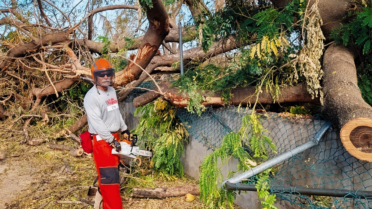 Team Rubicon Greyshirt Don Aiken holds a chainsaw in front of massive trees felled by Hurricane Melissa while clearing space for a water tank in Jamaica. 152 characters — just over. Tighter: Greyshirt Don Aiken holds a chainsaw in front of trees felled by Hurricane Melissa while clearing space for a potable water tank in Jamaica. 139 characters