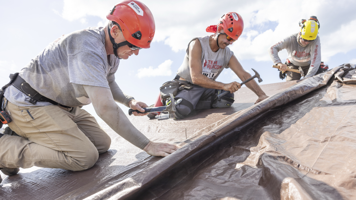 Three Team Rubicon volunteers in hard hats nail roof tarping onto a storm-damaged home in Jamaica.