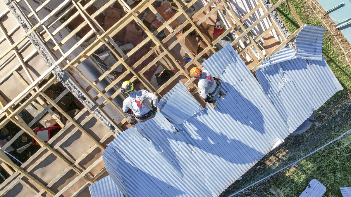 Aerial view of two Team Rubicon volunteers working on a severely storm-damaged roof with exposed rafters in Jamaica.