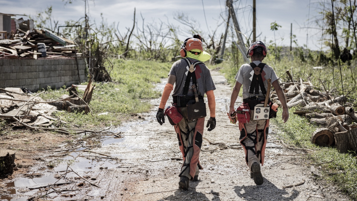 Two Team Rubicon volunteers in protective gear walk down a debris-strewn road through a storm-devastated neighborhood in western Jamaica.