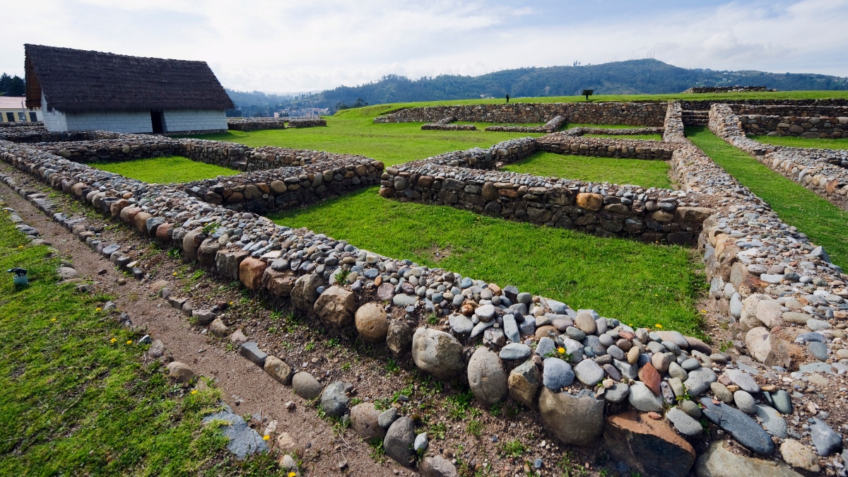 Stone foundation walls of the Inca ruins at Ingapirca, Ecuador, set against green hillside landscape under cloudy skies.