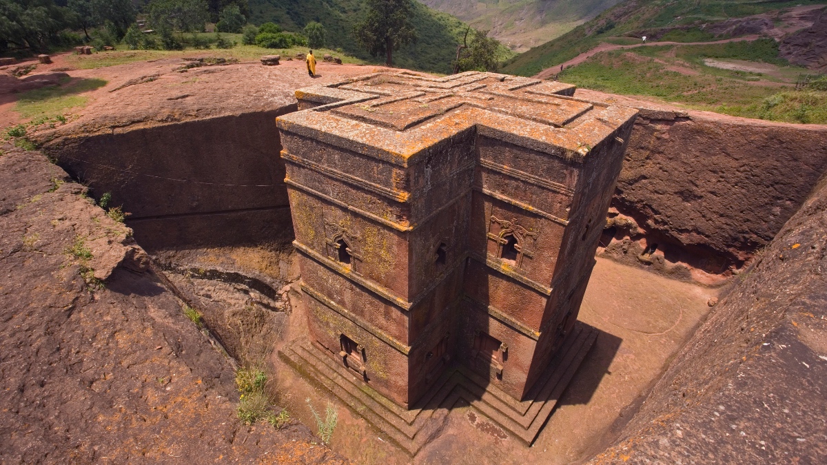 Aerial view of Bete Giyorgis, a cross-shaped rock-hewn church carved into red volcanic stone at Lalibela, Ethiopia, with a lone figure standing at the edge.