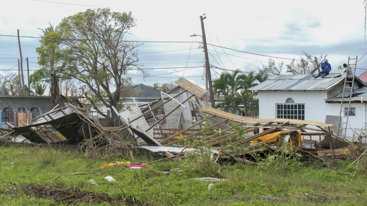 Workers on a ladder repairing a roof beside a collapsed structure in a Jamaican neighborhood after Hurricane Melissa.