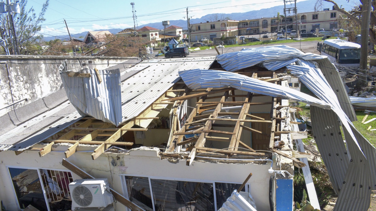 Street-level view of a house with roof torn open and metal sheeting buckled, with damaged buildings and mountains visible behind.