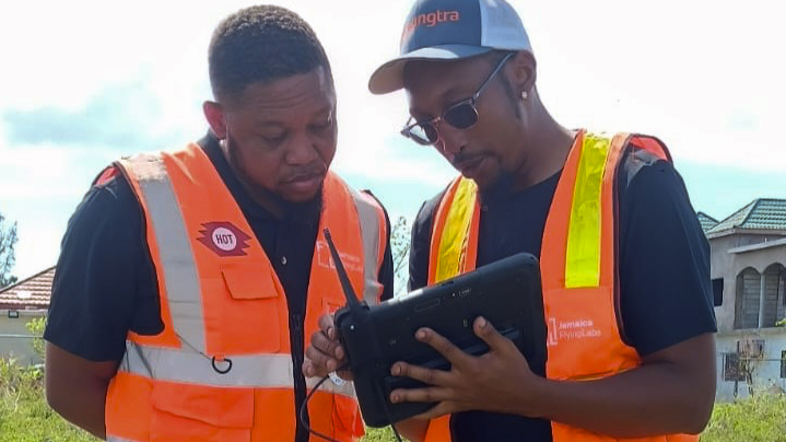 A HOT volunteer and Jamaica Flying Labs pilot in safety vests review a tablet together in the field after Hurricane Melissa.