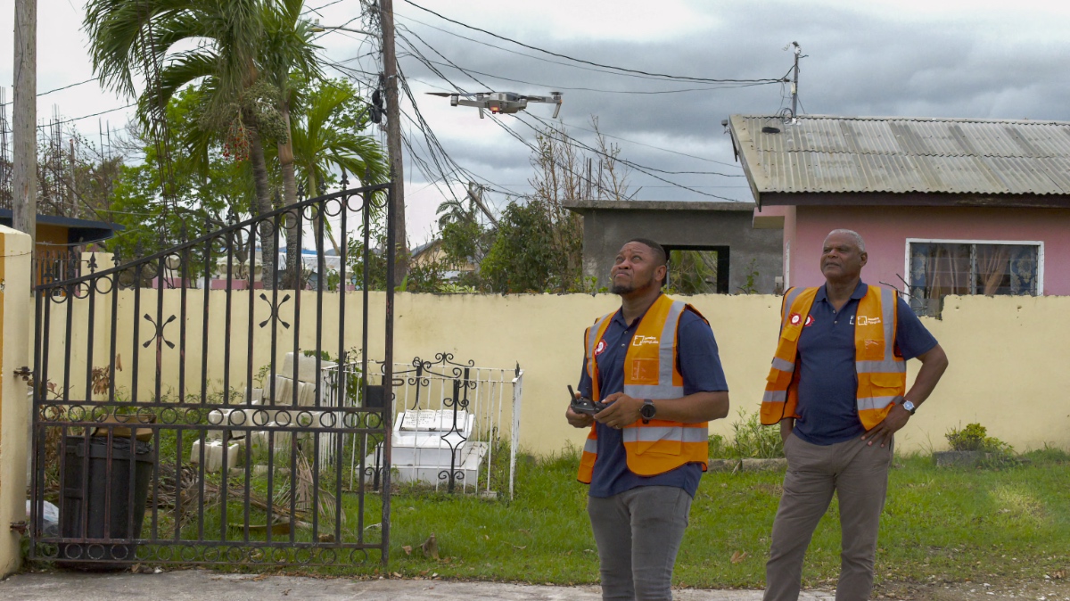 Two Jamaica Flying Labs team members in orange safety vests watch a drone rise above a hurricane-damaged Jamaican neighborhood.