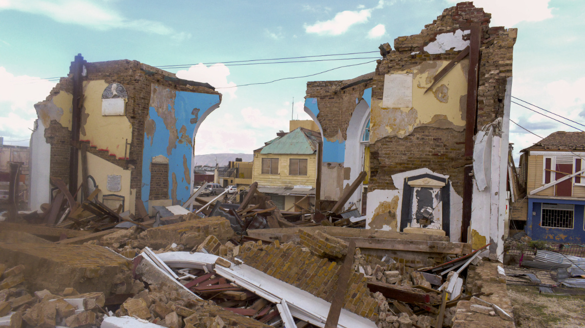 Collapsed brick building with walls and arches standing amid rubble in a Jamaican neighborhood after Hurricane Melissa.