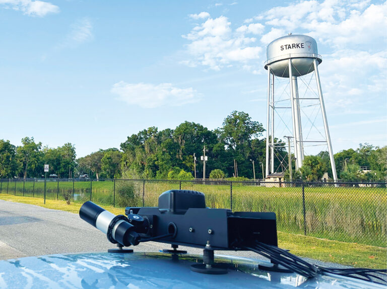 Black and silver technical equipment mounted on a vehicle, with a large silver water tower labeled "Starke" in the background under a partly cloudy sky.