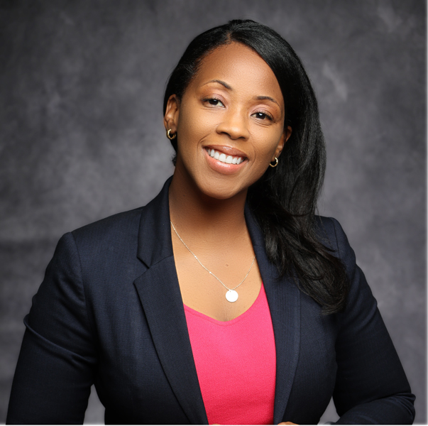 A headshot of a smiling woman with dark hair, wearing a pink shirt with a black blazer and gold necklace, posing against a gray background.