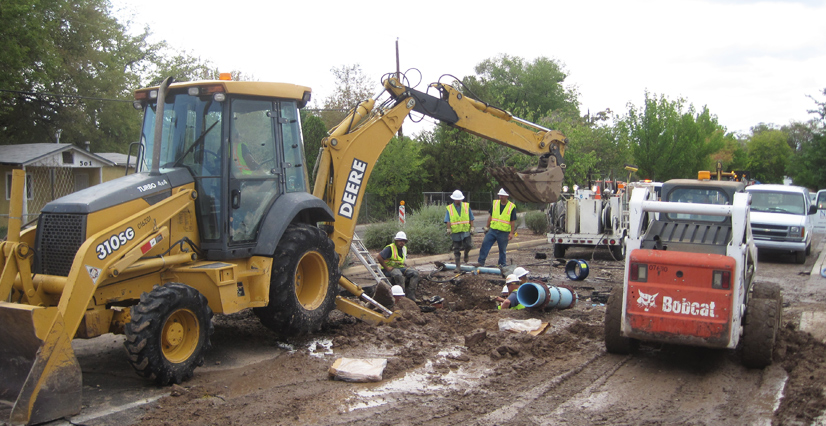 Construction workers in hard hats and safety vests install a large blue pipe in a muddy ditch, with a yellow backhoe and a white Bobcat skid-steer loader nearby.