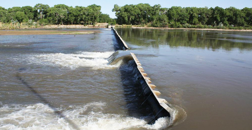A low dam spans a wide river, with white water flowing over its edge. Dense green trees line the far riverbank.