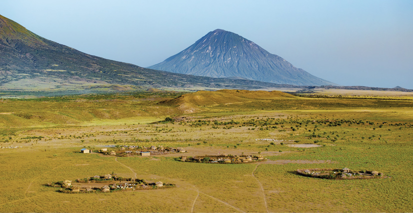 Four small circular villages with thatched huts dot a wide grassy plain, with a large cone-shaped volcano in the background.