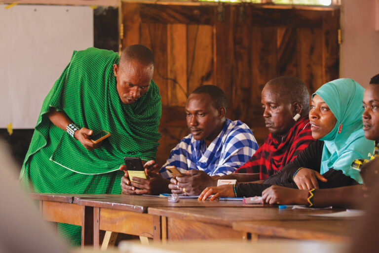 A man in a green patterned outfit stands and leans over a seated man as they look at a smartphone together in a classroom. Other men and a woman in a blue hijab sit beside them at a wooden desk.
