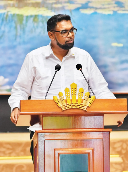 A bearded man with glasses in a white shirt speaks at a wooden podium with a gold emblem and two microphones.