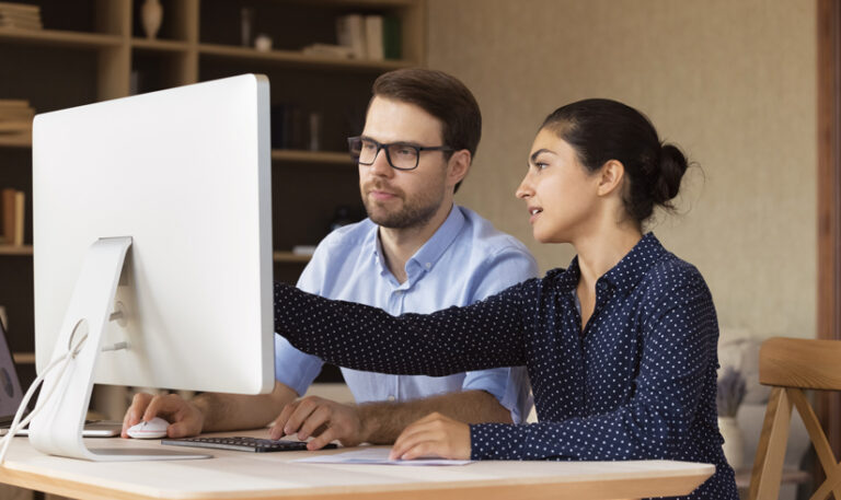 A woman points at a computer screen while a man sits beside her, both collaborating at a desk.