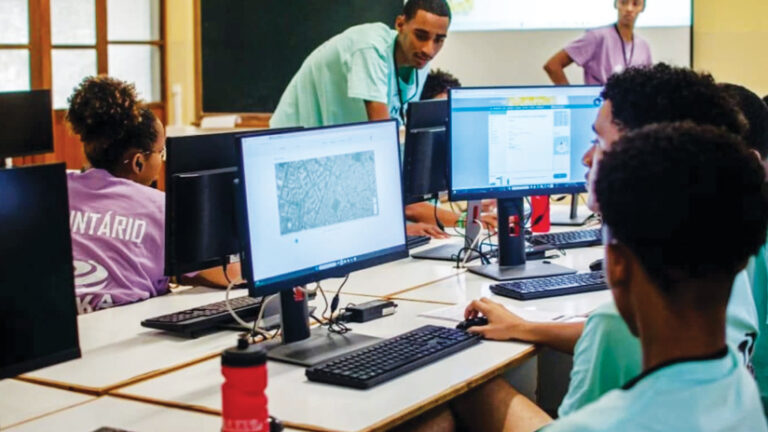 In a computer lab, an instructor leans over to help a student at a desktop computer, while other students work at separate stations.