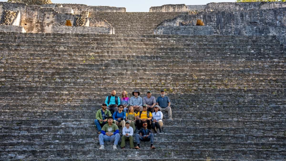 A group of approximately fourteen researchers and archaeologists sit together on the broad limestone steps of a towering Maya pyramid, dwarfed by the massive stone staircase rising behind them into an overcast sky.