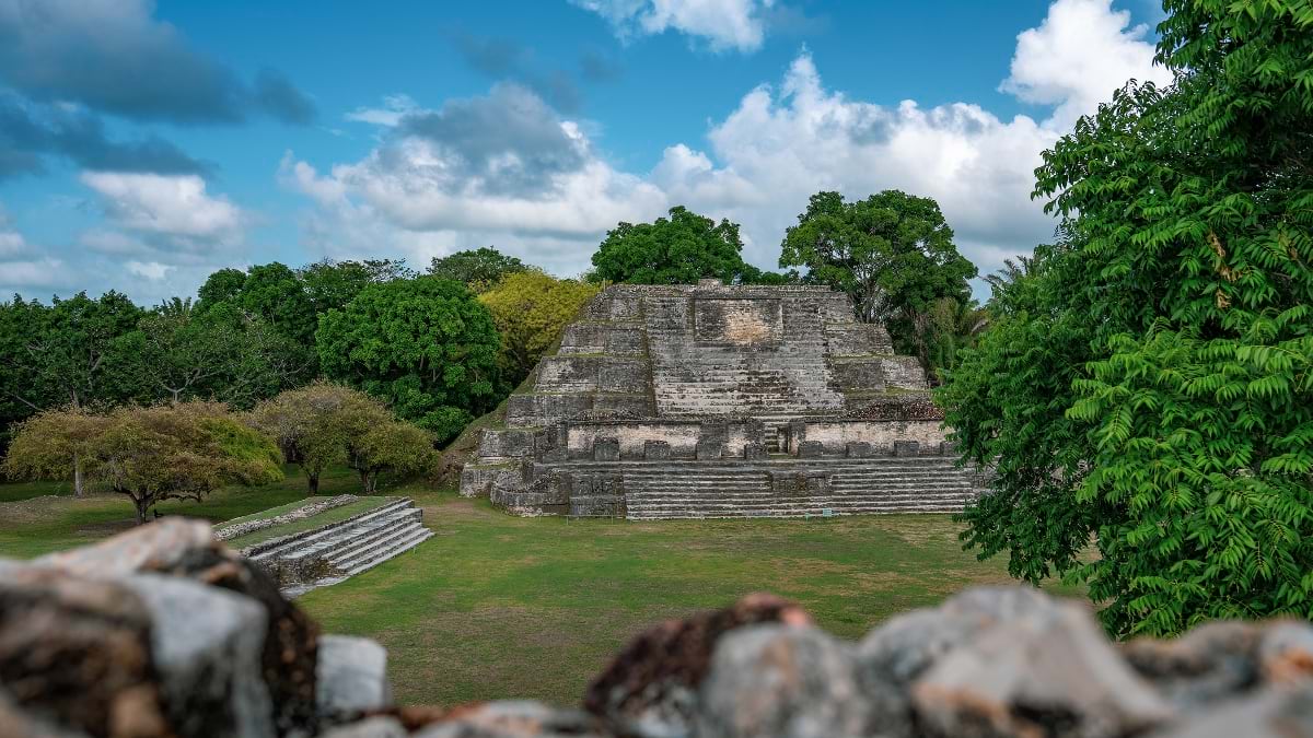 A wide-angle view of a large stepped Maya pyramid at Altun Ha, photographed from an elevated position across a green plaza, with blurred stone ruins in the foreground and dense tropical trees flanking the structure under a partly cloudy blue sky.