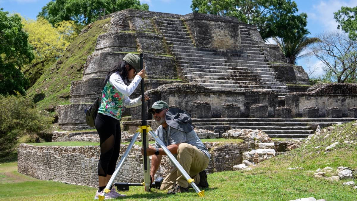 Two researchers set up a survey tripod on the grass in front of a stepped Maya pyramid. A woman in a floral vest adjusts the instrument from above while a man crouches at the base to level it.