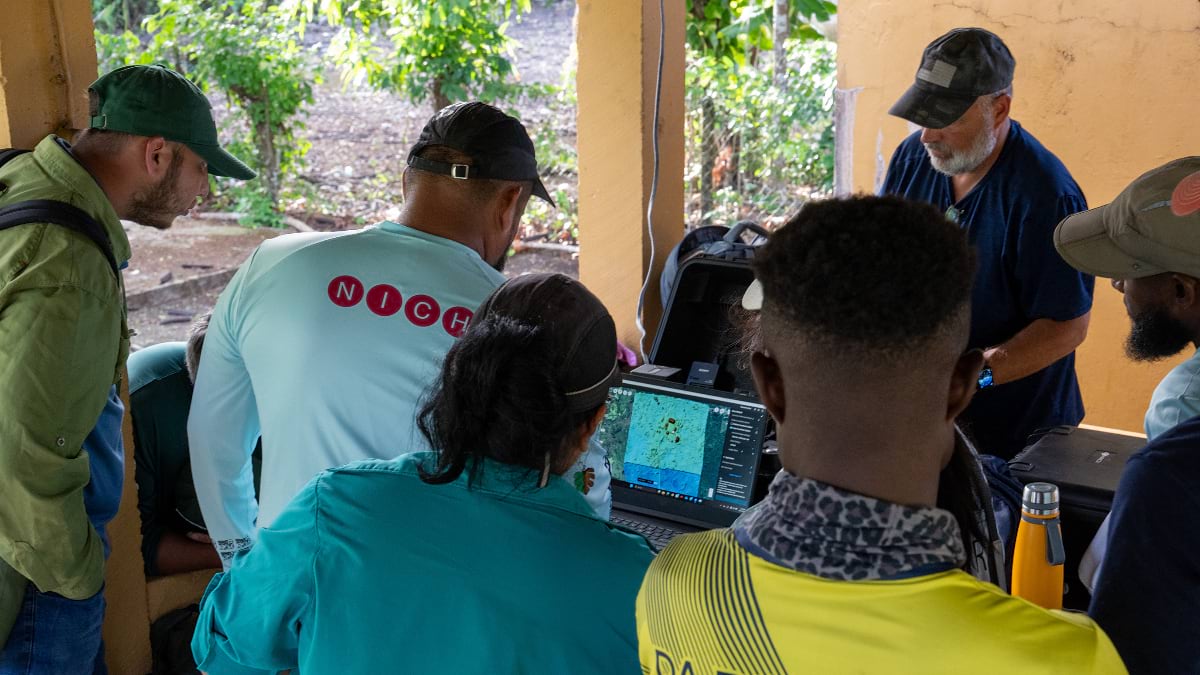 A group of five researchers crowd around a laptop displaying an aerial map, gathered in an open-air structure with a yellow wall and jungle visible behind them. One person's shirt bears the NICH logo.