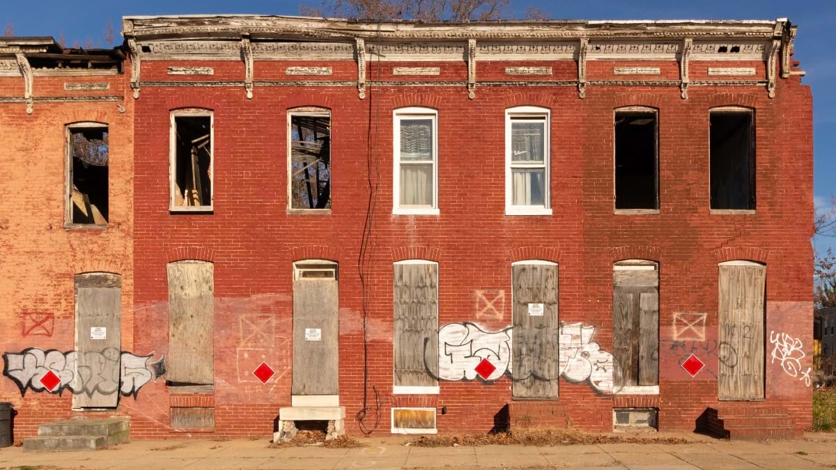 A row of condemned Baltimore brick row houses with boarded doors, graffiti, and red diamond hazard markers on the facades.