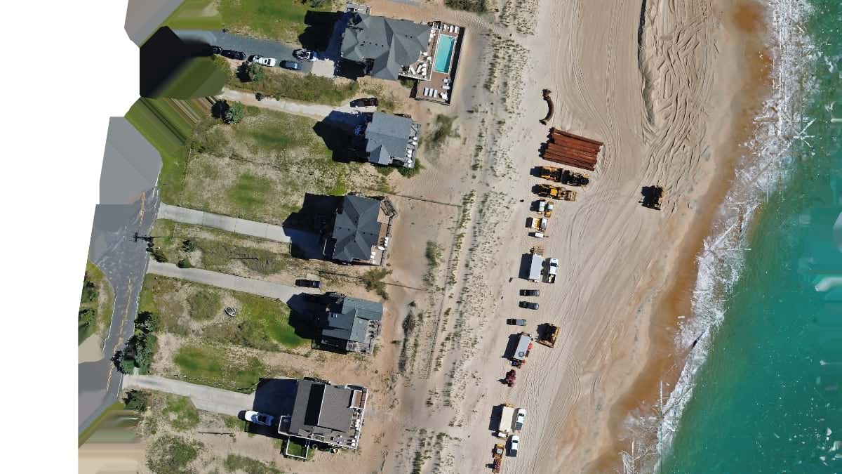 Aerial drone view of beach nourishment equipment and pipe staged on Outer Banks shoreline beside oceanfront homes.