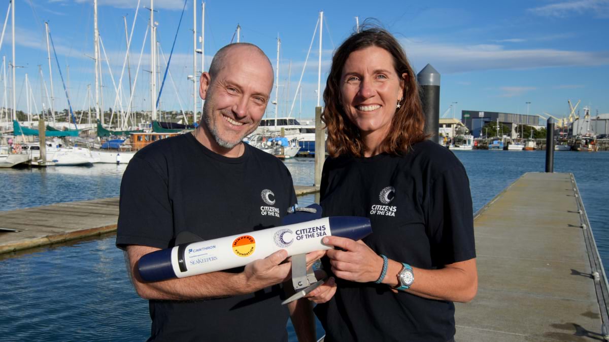 Two people in Citizens of the Sea t-shirts hold a TorpeDNA sampler on a marina dock with sailboat masts behind them.