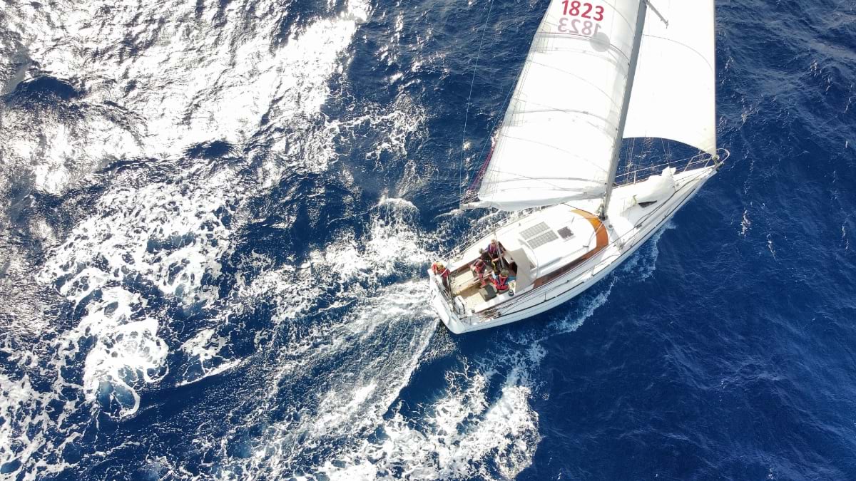 Aerial view looking directly down at a white-hulled sailboat under full sail, sail number 1823 visible, cutting through deep blue Pacific water with wake fanning out behind the hull. Two crew members are visible in the cockpit.