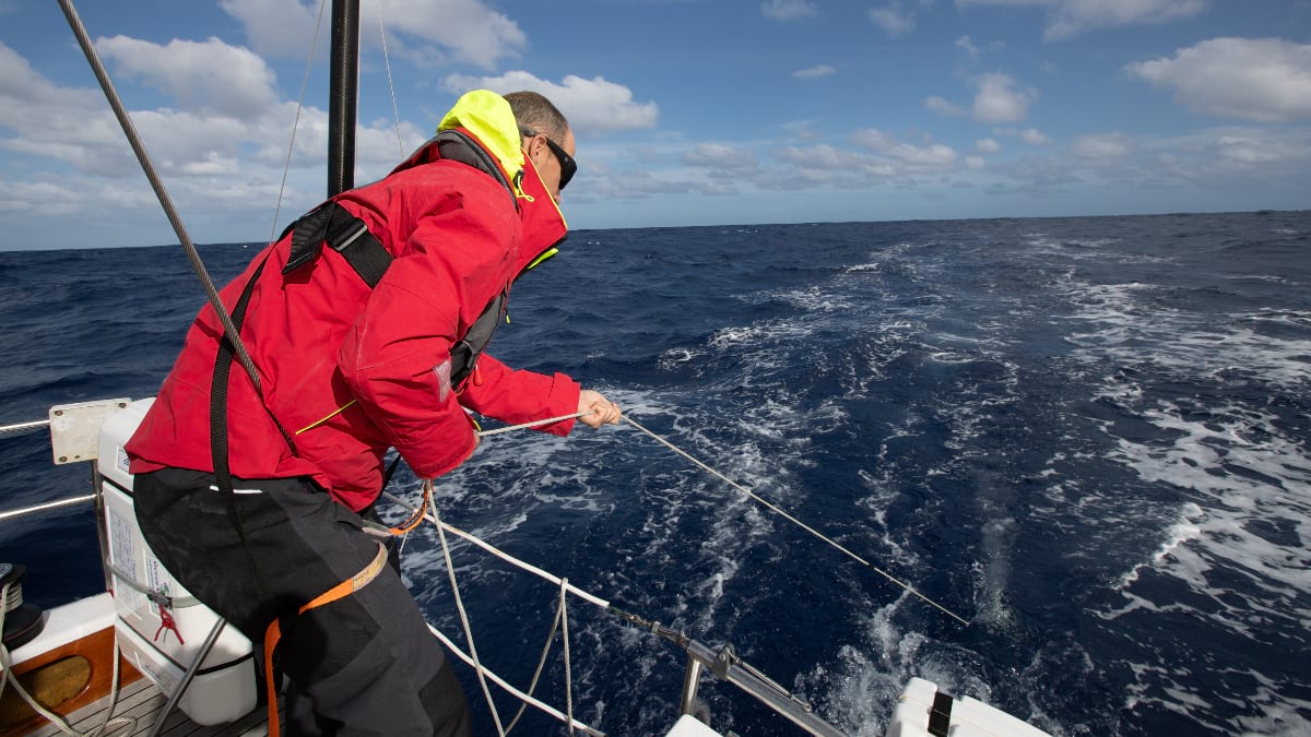 A person in red foul-weather gear and a safety harness leans over the stern rail of a moving sailboat to pull in the TorpeDNA sampler by its tether line. The open Pacific stretches to the horizon under a partly cloudy sky.