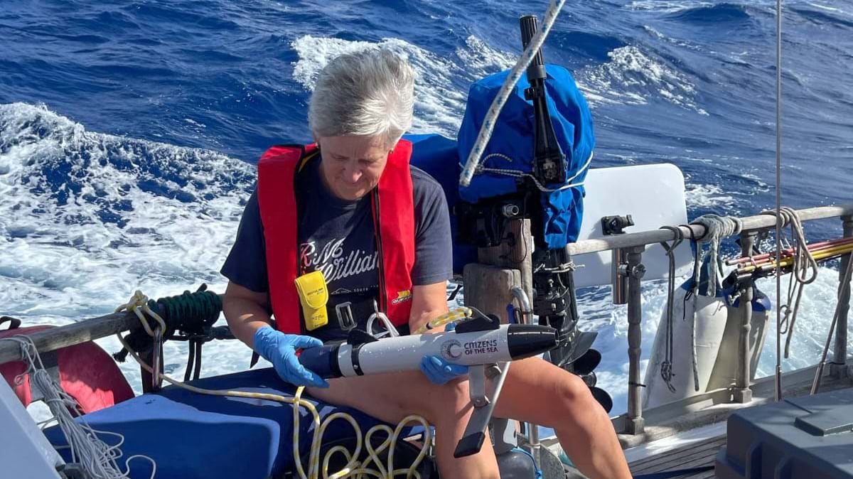 A woman in a red inflatable life vest and blue nitrile gloves handles a white torpedo-shaped sampling device marked "Citizens of the Sea," seated at the stern of a moving vessel with open blue water behind her.