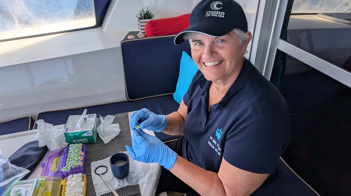A woman wearing a Citizens of the Sea cap and blue nitrile gloves smiles at the camera while handling a small circular filter at a cabin table. Sample tubes, Kimtech wipes, and sample trays are spread across the surface in front of her.