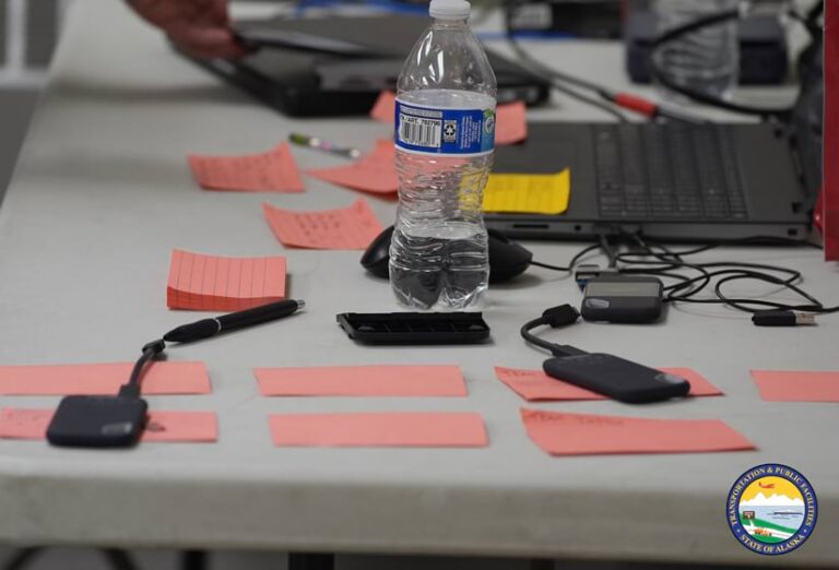 Photo of a while folding table covered by pink sticky notes, laptops, black hard drives, and a half-full water bottle.