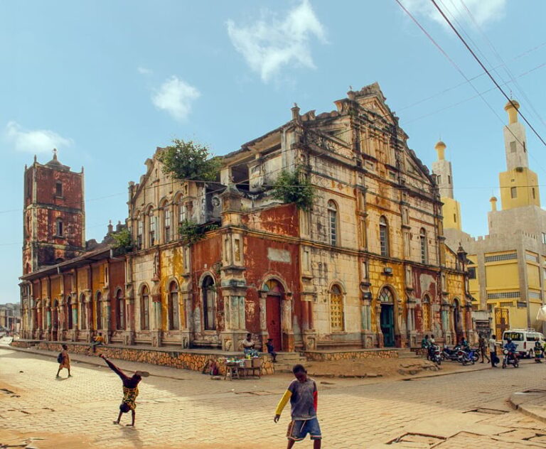 Photo of a large church with discolored paint and vegetation sprouting from the upper stories Children are playing in the street in front of the building.