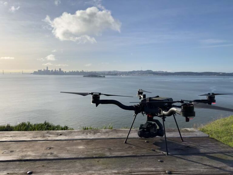 Photo of a drone perched on a park bench in front of a body of water with a city skyline in the bakground.