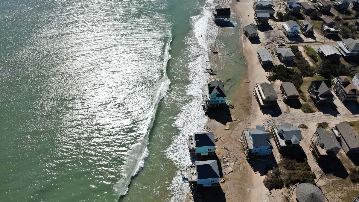 Nadir drone view of three Buxton homes in the surf mid-collapse, with debris on the beach and sandbags visible at the frame's edge.