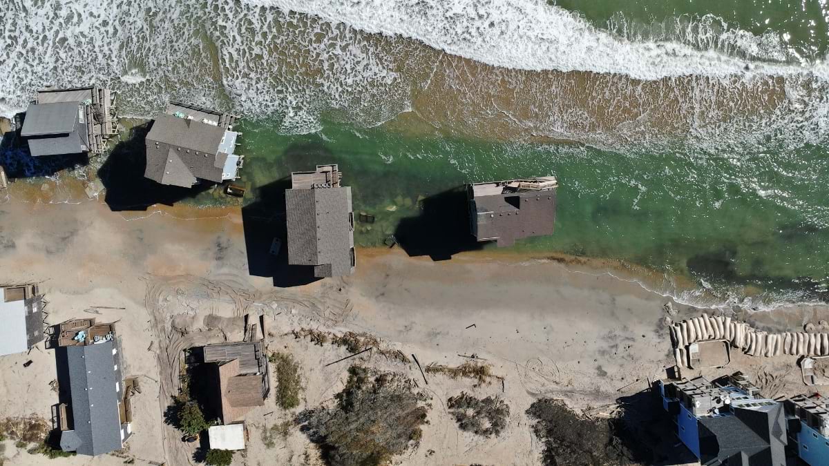 Nadir drone view of a collapsed Buxton home in the surf surrounded by debris, displaced sand, and overwhelmed sandbags.