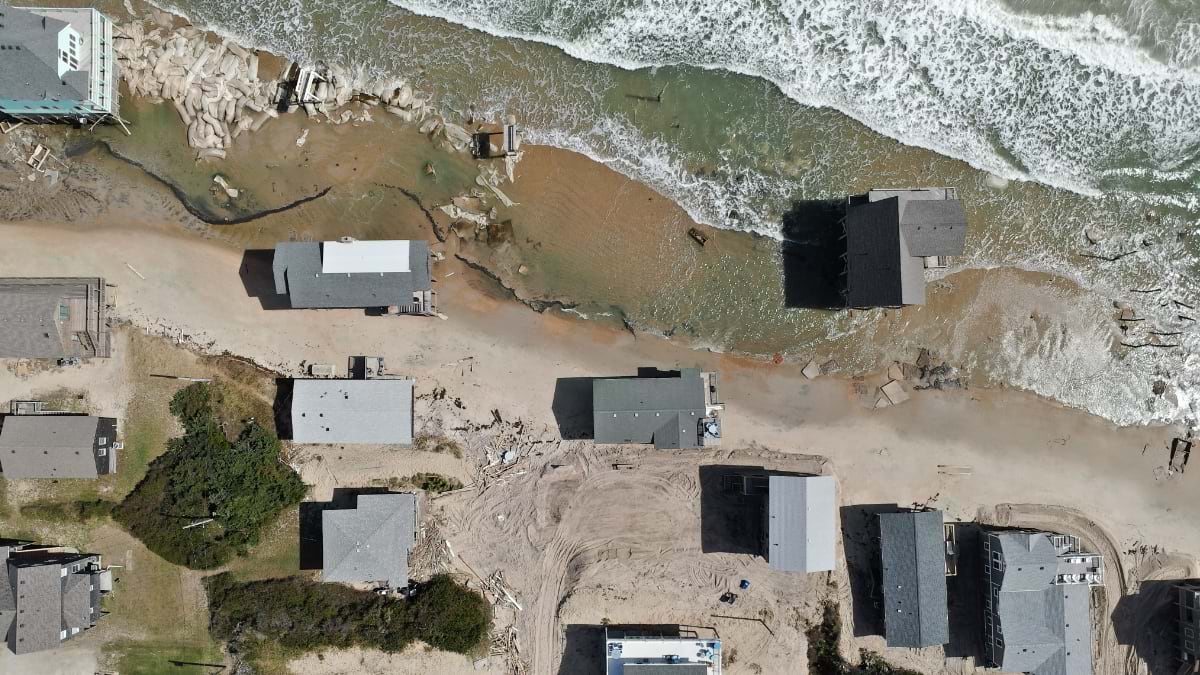 Oblique drone view of a Buxton sandbag line shielding a row of homes from the surf, with one stilt structure already standing in the water.