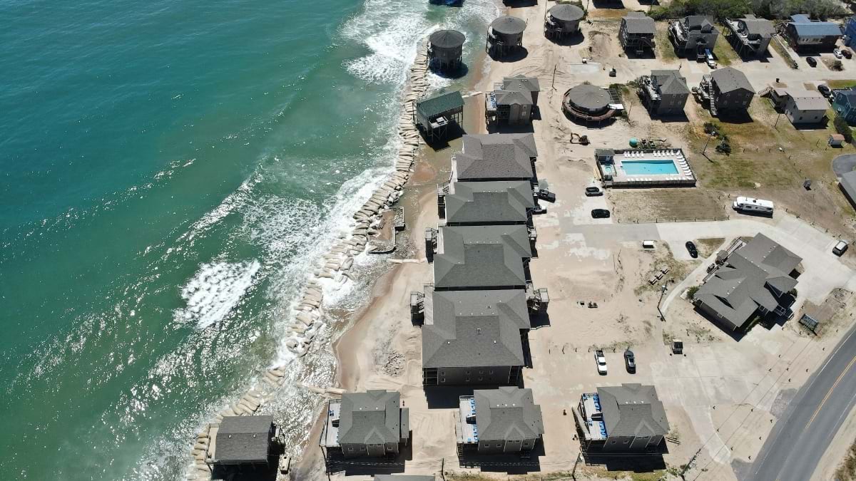 Oblique drone view of Buxton showing a row of homes at the waterline with no beach buffer remaining and waves breaking against foundations.