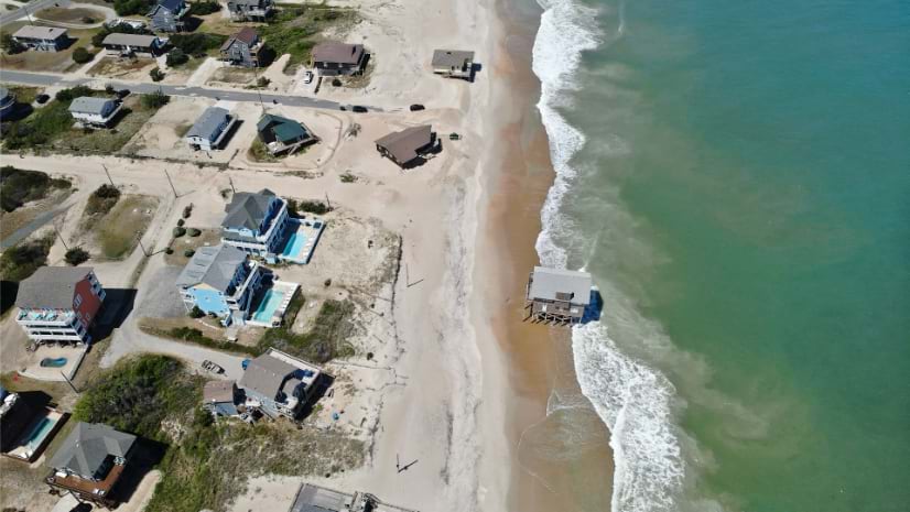 Oblique drone view of Rodanthe showing a stilt house in the surf and a narrow strip of beach separating remaining homes from the Atlantic.