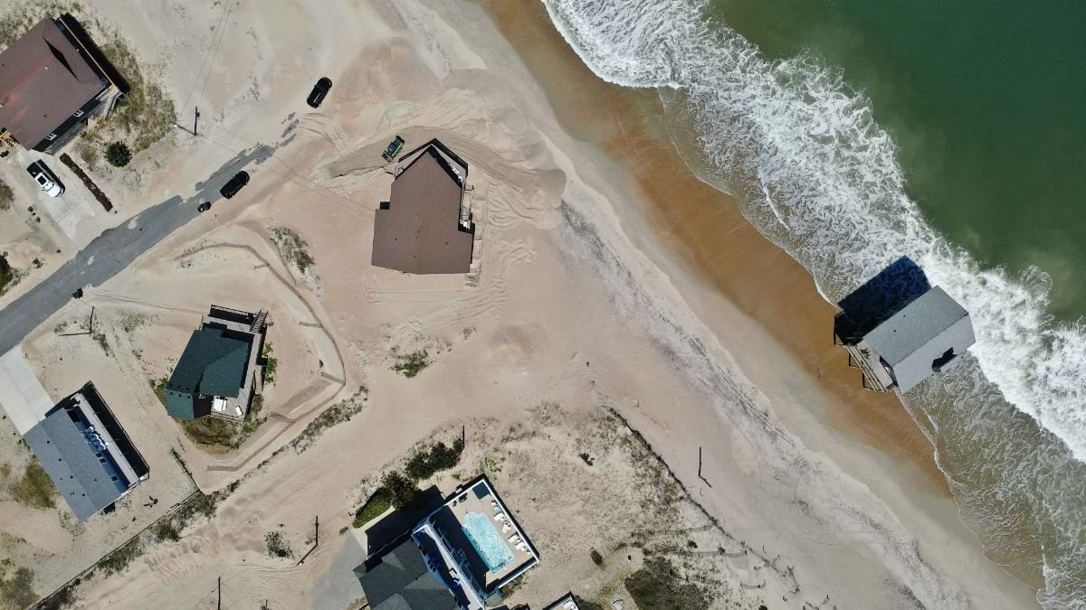 Nadir drone view of a Rodanthe house stranded on bare sand as waves reach a neighboring home already in the surf.