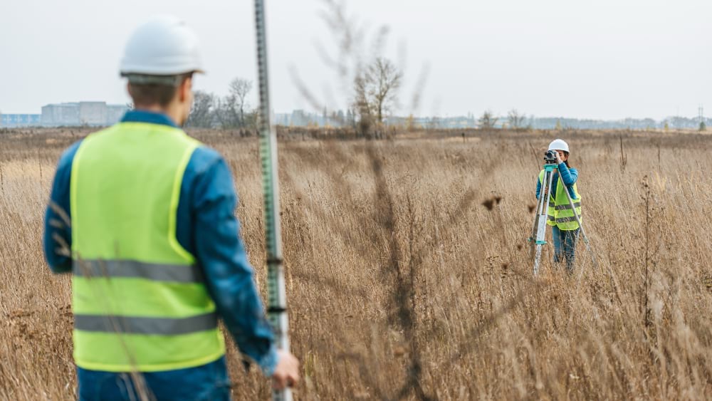 Two surveyors in high-visibility vests working across a dry grass field, one holding a rod while the other operates a theodolite.
