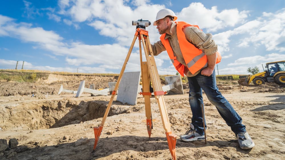 Surveyor in orange vest operating a theodolite on an active construction site with excavation equipment visible in the background.