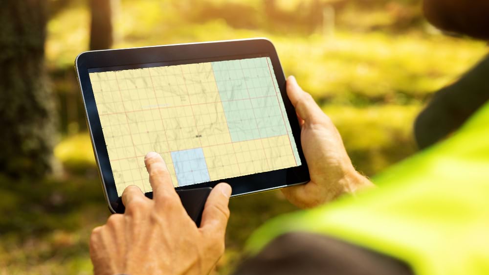 Field worker in high-visibility vest holding a tablet displaying a topographic map in a forested outdoor setting.