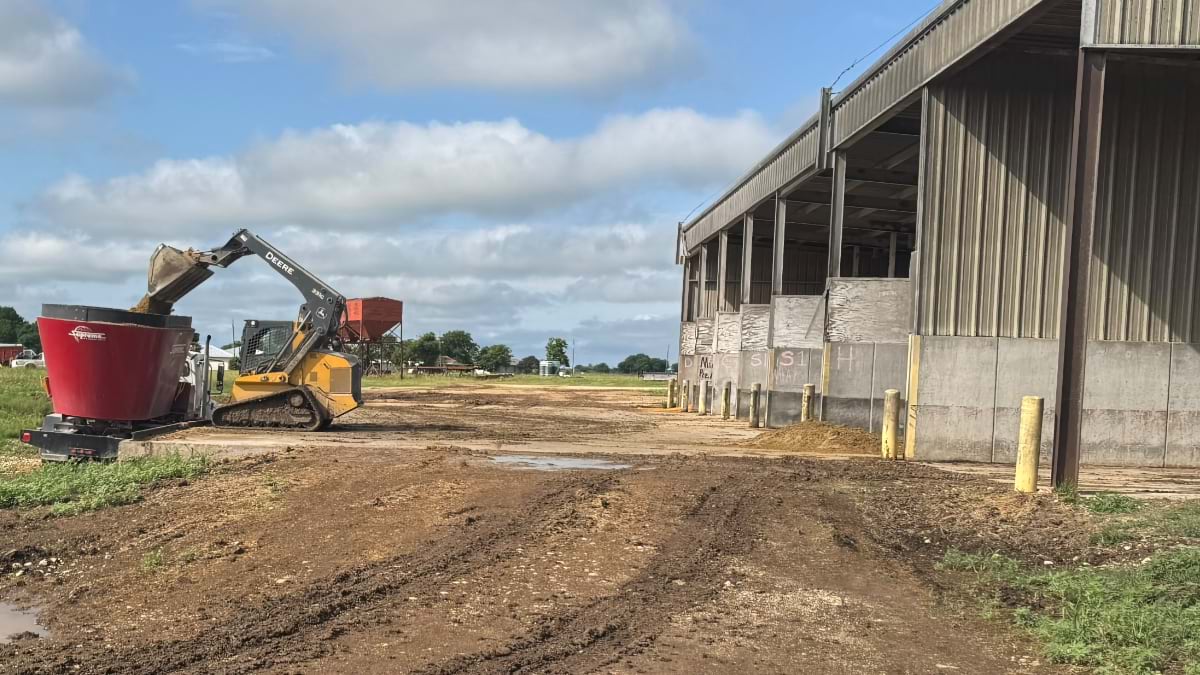 John Deere skid steer loading a red feed wagon outside a feedlot barn at McGregor Research Center.