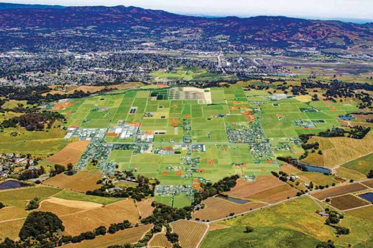 Oblique aerial view of a valley with agricultural fields and vineyards in the foreground, a town at the base of mountains in the background, and a large digitally rendered map overlay with parcels of land outlined in red.