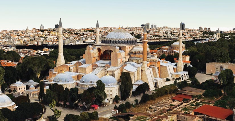 An oblique aerial 3D rendition of the Hagia Sophia in Istanbul, showing its large central dome, smaller domes, and four minarets. The building is surrounded by trees, with cityscape in the background and archaeological ruins in the foreground.